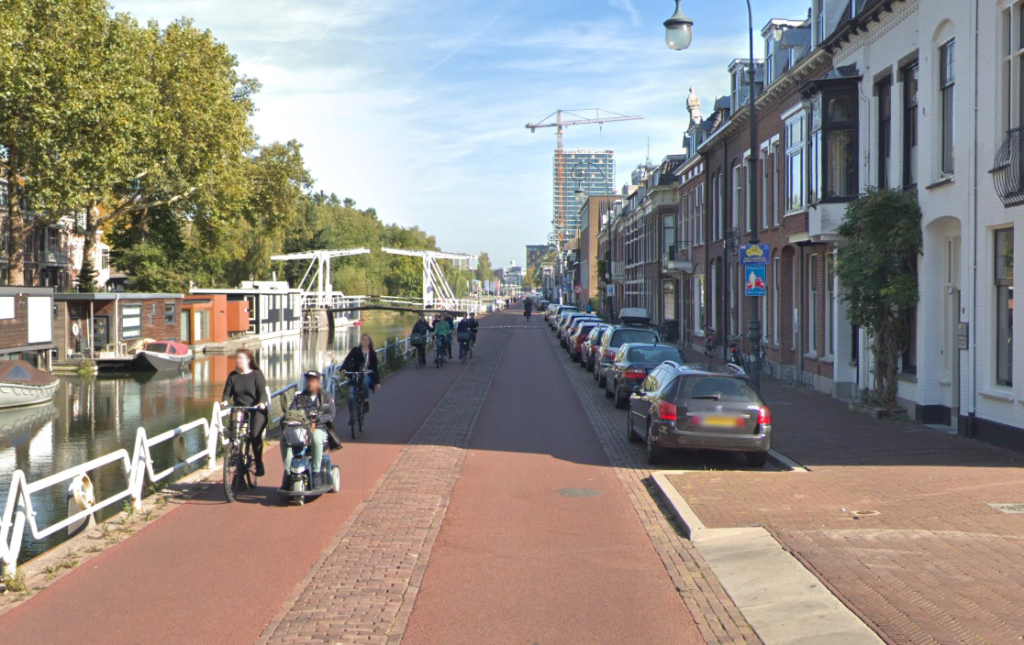 Imagen de la Leidseweg de Utrecht. Es una imagen de una calle que transcurre paralela a un canal, que se encuentra en el lado izquierdo. La calzada se compone de dos bandas de asfalto rojo, como si fueran dos carriles bici, separadas entre si por una banda de adoquines. A la derecha de la calzada, se encuentra una banda de coches aparcados y una acera paralela a una fila de casas de dos plantas.

En la calle se observan badenes para reducir el tráfico, así como a en torno una docena de personas circulando en bicicleta y en vehículos de mobilidad personal.